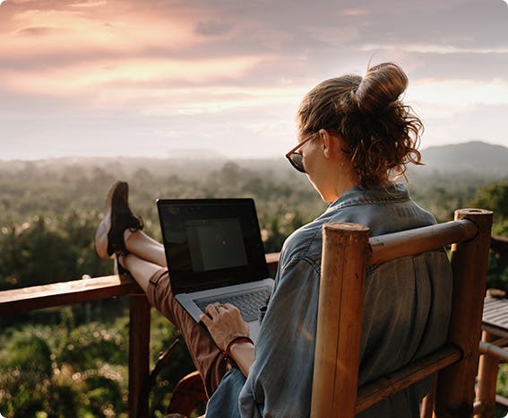A woman sitting on a balcony with a scenic view while working on her laptop